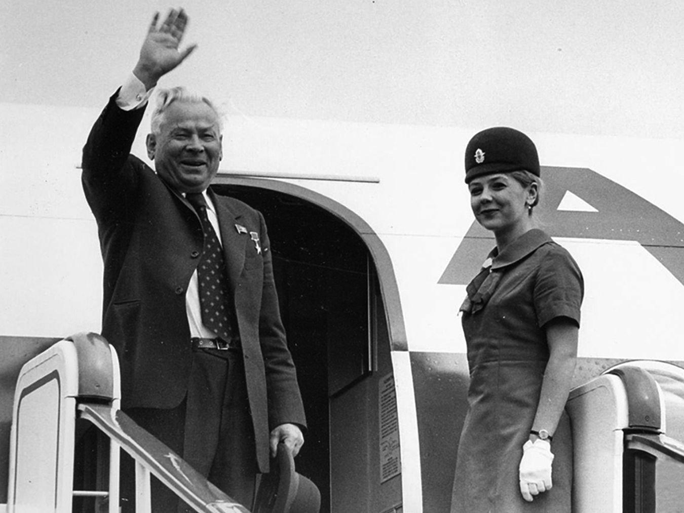 Image of smiling man waving farewell as he boards a plane, a stewardess at his side.